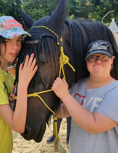 Zwei lächelnde Jugendliche stehen eng beieinander mit einem Pferd in der Mitte, wobei beide es mit den Händen berühren. Das Pferd trägt eine schwarze Fliegenmütze und eine gelbes Geschirr, und die Jugendlichen tragen jeweils eine Kappe.