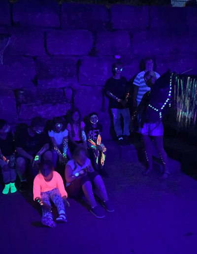 Eine Gruppe von etwa zwölf Menschen, darunter mehrere Kinder, sitzt und steht im Dunkeln vor einer Steinmauer, wobei ihre Kleidung und Körperbemalung durch Schwarzlicht in leuchtenden Neonfarben strahlt.