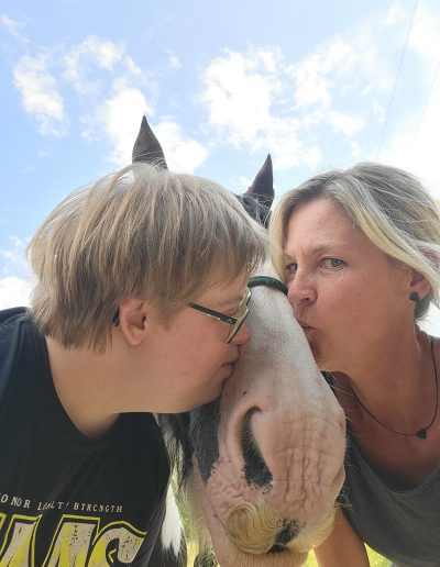 Ein Kind und eine erwachsene Frau küssen gleichzeitig ein Pferd, das im Zentrum des Bildes steht. Das Kind und die Frau neigen sich von links und rechts dem Pferdekopf zu, während sie unter einem blauen Himmel im Freien stehen.