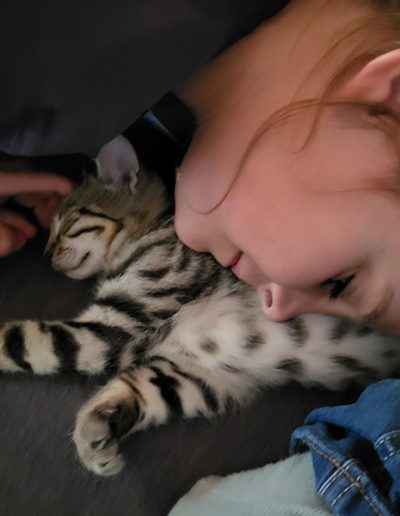 Eine junge Person liegt eng an ein schlafendes, gestromtes Kätzchen geschmiegt, das entspannt auf einer dunklen Decke oder Kleidung liegt.