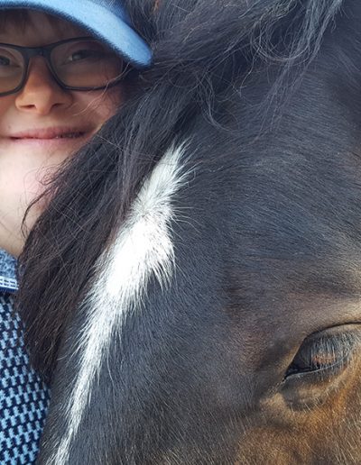 Eine lächelnde Person mit Brille und blauer Baseballkappe schmiegt sich eng an den Kopf eines dunkelbraunen Pferdes.