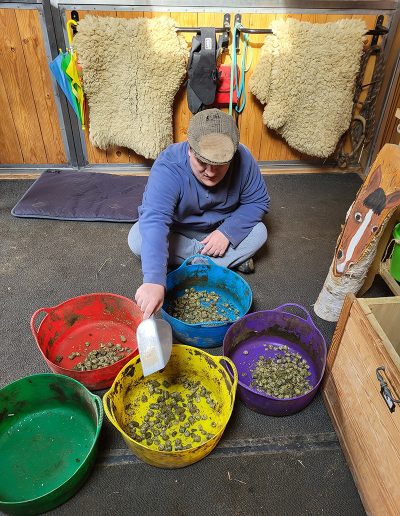 Ein Mann mit einer braunen Schiebermütze sitzt in einem Stall auf dem Boden und verteilt Pferdefutter mit einer weißen Schaufel in fünf großen, bunten Eimern. Im Hintergrund hängen zwei helle, flauschige Lammfelle an einer Holzwand.