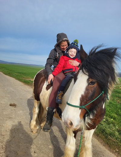 Eine erwachsene Person reitet auf einem großen, braun-weiß gescheckten Pferd und hält ein kleines Kind im roten Anorak vor sich im Sattel. Beide lächeln fröhlich in die Kamera, während das Pferd auf einem Feldweg unter weitem blauem Himmel steht.