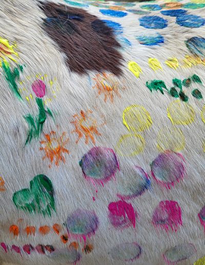 Das Bild ist eine Nahaufnahme des hellen Fells eines Pferdes, das vollständig mit bunten Motiven wie Herzen, Blumen und Punkten bemalt ist.