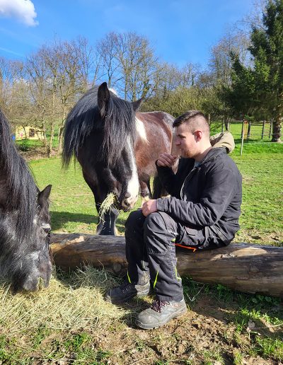 Ein Mann sitzt im Freien auf einem liegenden Baumstamm und blickt nachdenklich zur Seite.