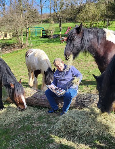 Ein Mann sitzt im Freien auf einem Baumstamm, umgeben von vier großen Pferden, die alle Heu fressen, das auf dem Boden liegt. Die Szene findet auf einer grünen, leicht abfallenden Wiese mit Bäumen und einem Spielgerät im Hintergrund unter blauem Himmel statt.