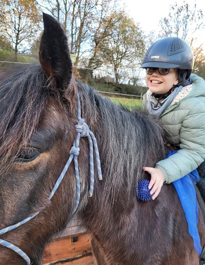Ein Mädchen mit Reithelm und Brille sitzt lächelnd auf einem dunklen Pferd und hält eine blauen Igelball in der Hand, die auf dem Pferdehals liegt. Im Hintergrund sind Bäume und eine Wiese bei mildem Abendlicht zu sehen.