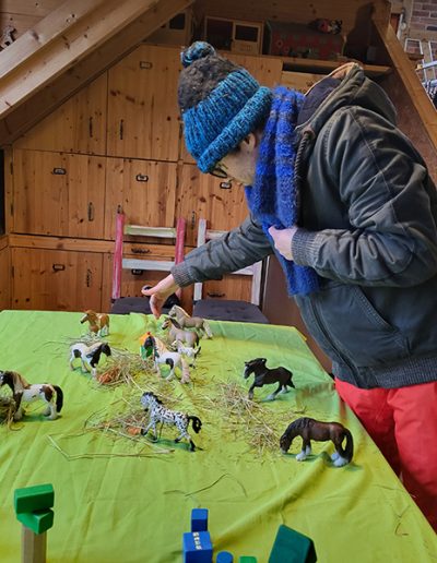 Hier ist die Bildbeschreibung: Eine Person mit blauer Mütze und Schal beugt sich über einen Tisch, auf dem sie eine Szene mit vielen kleinen Spielzeugpferden, Heu und Bauklötzen arrangiert hat. Die Szene findet in einem gemütlichen Raum mit Holzwänden und einer Holztreppe statt.