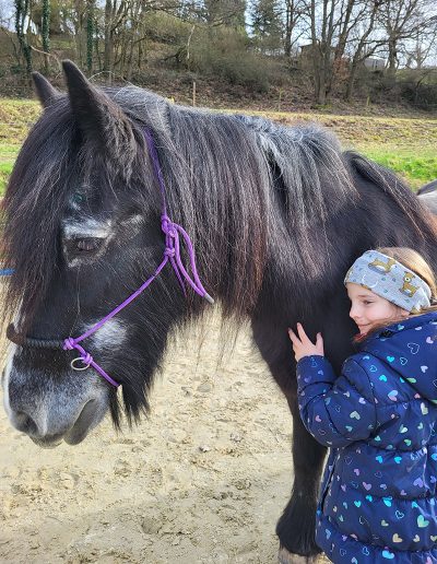 Ein Mädchen mit einer dunklen Jacke und einem Stirnband steht lächelnd auf einem Sandboden