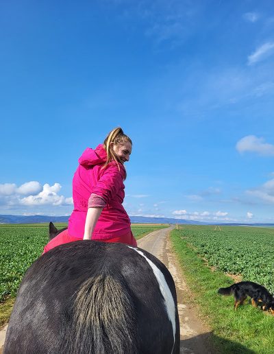Eine lächelnde Frau in einer leuchtend pinken Jacke reitet auf einem dunklen Pferd einen Feldweg entlang.