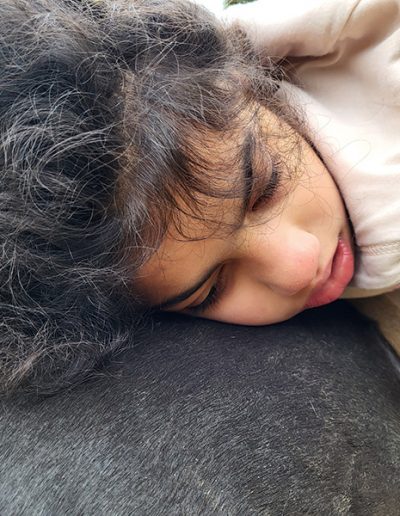 Ein Mädchen mit dunklen Locken und einer gelben Haarspange liegt entspannt auf dem dunklen Rücken eines Pferdes. Es trägt einen beigen Kapuzenpullover, und die Szene strahlt Ruhe und Vertrauen aus.