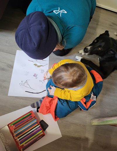 Eine erwachsene Person und ein Kleinkind sitzen gemeinsam auf dem Boden und schauen auf eine Zeichnung, während sie bunte Stifte in einer Schachtel vor sich haben.