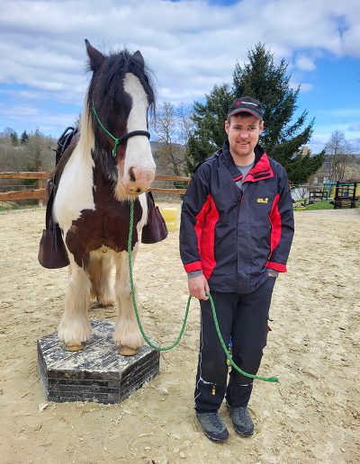 Ein Mann in einer rot-schwarzen Jacke steht lächelnd neben einem braun-weiß gescheckten Pferd, das auf einer erhöhten schwarzen Plattform steht.