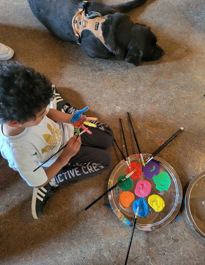 Ein Kind sitzt auf dem Boden und bemalt seine Hand mit leuchtend blauer Farbe, wobei es einen Pinsel benutzt und eine Palette mit bunten Farbtupfern vor sich hat.