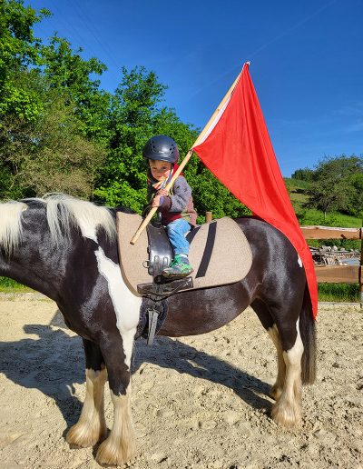 Ein kleines Kind sitzt mit einem Reithelm und einem leuchtend roten Fähnchen auf einem schwarz-weiß gefleckten Pferd.