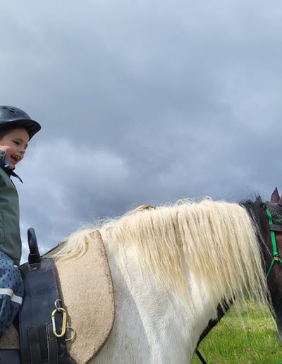 Ein kleines Kind mit Reithelm sitzt lächelnd auf dem Rücken eines braun-weißen Pferdes mit heller Mähne, unter einem dramatisch bewölkten Himmel.