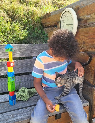 Ein Kind mit lockigem Haar sitzt auf einer Holzbank und streichelt eine grau-getigerte Katze, die auf seinem Schoß steht. Neben dem Kind befindet sich ein hoher Turm aus bunten Bauklötzen.