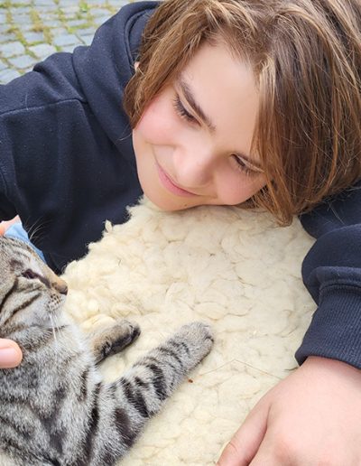 Ein lächelndes Kind liegt auf einem cremefarbenen Schaffell und streichelt sanft ein graubraun getigertes Kätzchen, das entspannt mit geschlossenen Augen daliegt